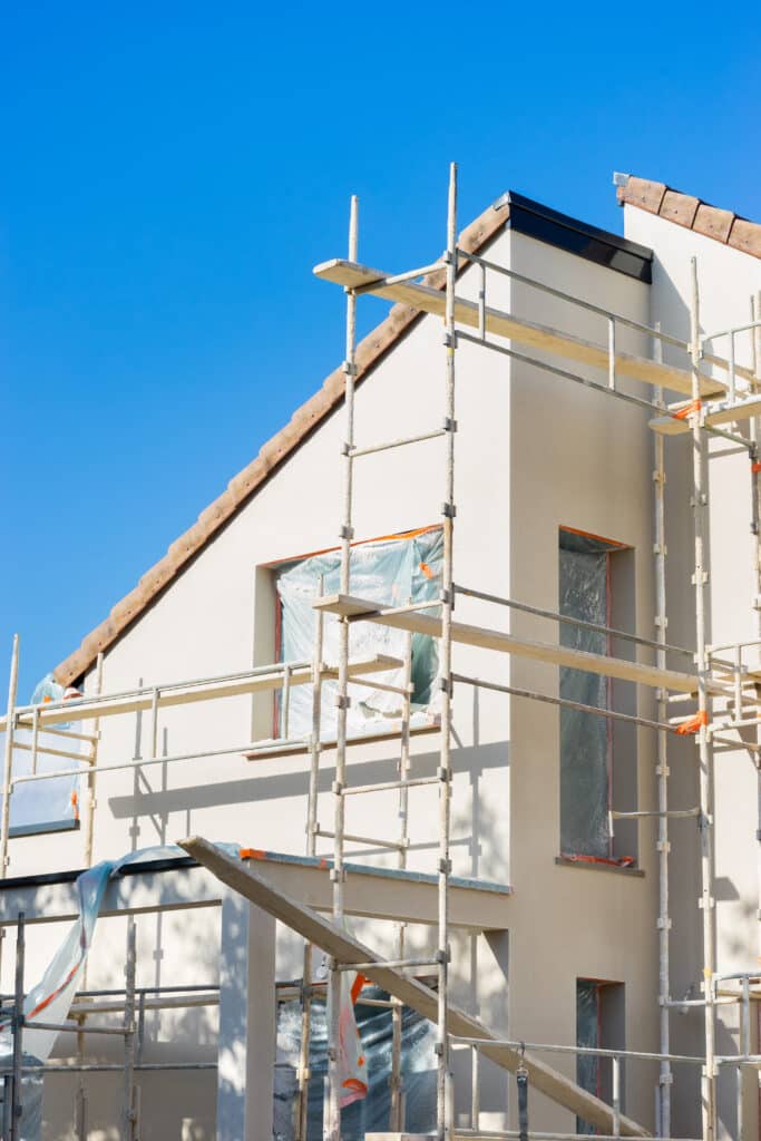 Maison neuve en construction avec enduit beige, échafaudage métallique et fenêtres bâchées sous un ciel bleu vif.
