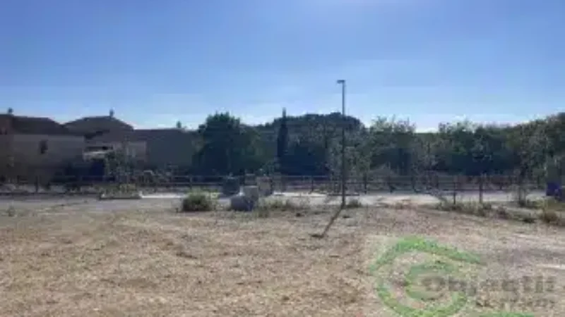 Terrain vague ensoleillé avec vue sur des maisons, arbres et un lampadaire, portant le logo Objectif terrains.
