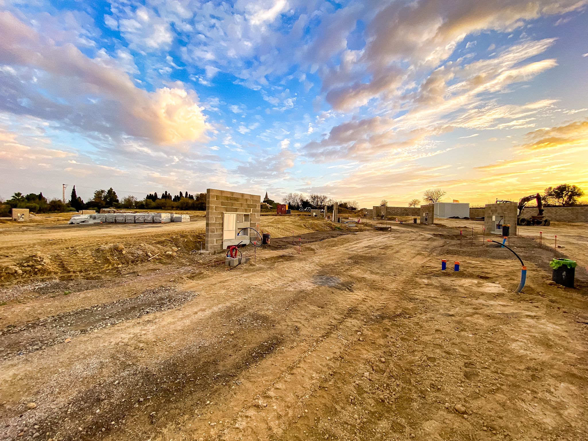 Chantier de construction en terre battue au coucher du soleil, murs en parpaings, ciel bleu et orange, pelle mécanique visible.
