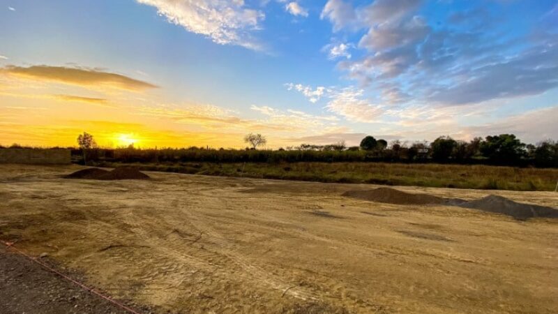 Vaste chantier de construction en terre battue au coucher du soleil, avec des tas de matériaux et une ligne rose de marquage.