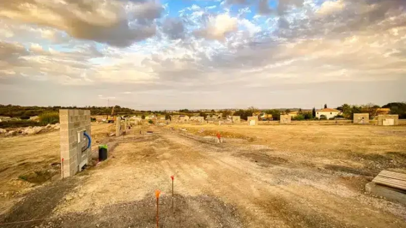 Chantier de lotissement en terre battue sous un ciel dramatique, blocs de béton et piquets de construction visibles.