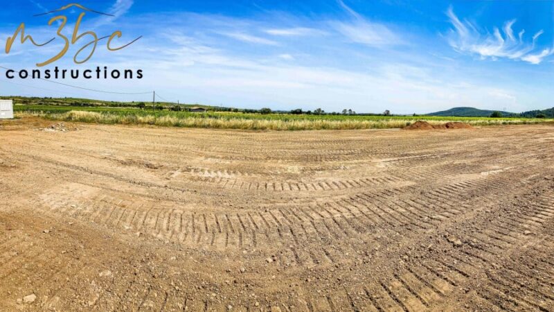 Terrain de construction M3BC Constructions, terre remuée avec traces de pneus sous un ciel bleu et nuageux, champ en arrière-plan.